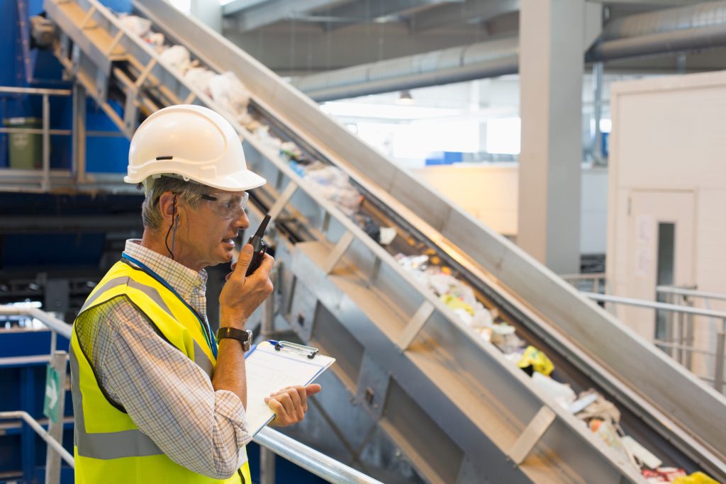 A man in a hard hat  and  safety vest speaking into a walkie talkie inside recycling facility.