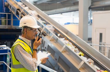 A man in a hard hat and safety vest speaking into a walkie talkie inside recycling facility.