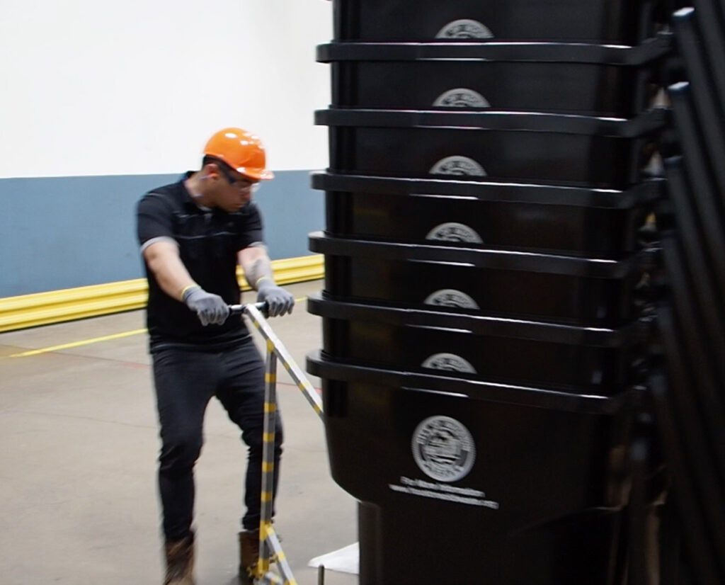 a man in an orange hard hat and black shirt moving a load of stacked recycling carts
