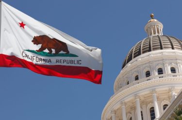 The California state flag featuring a grizzly bear and the words "California Republic" flutters against a clear blue sky, with the dome of the California State Capitol building visible in the background.