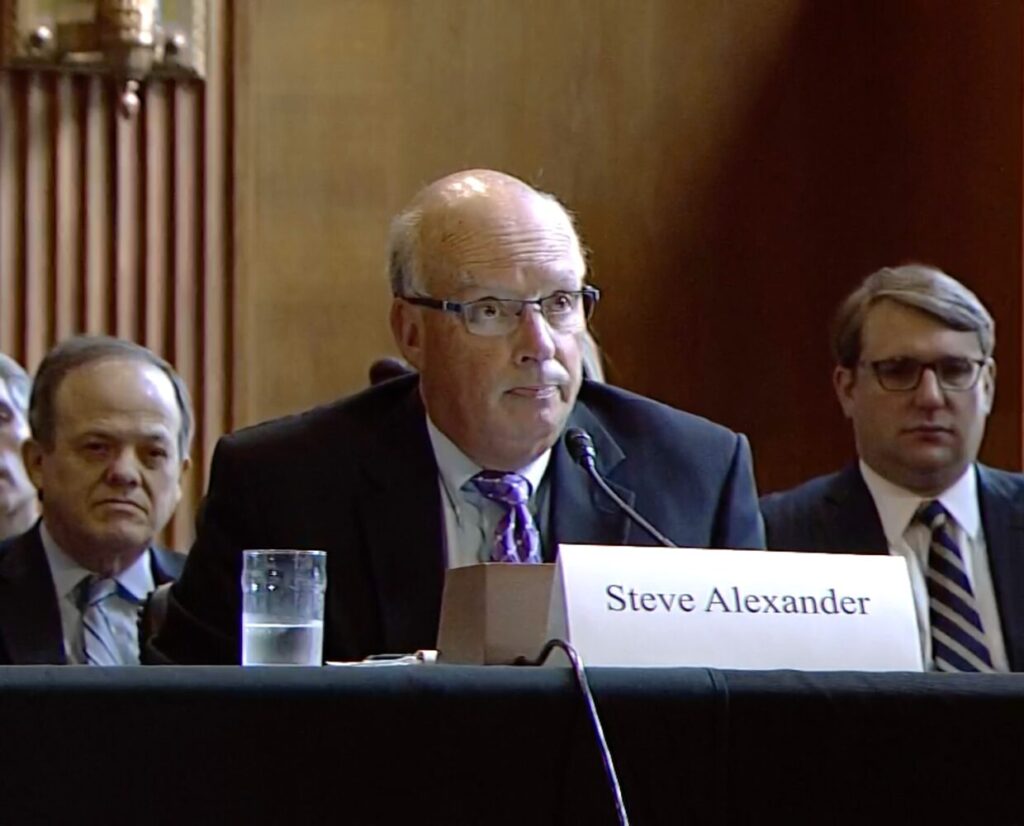 A man in a suit in front of a microphone with a plaque that states Steve Alexander