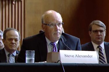 A man in a suit in front of a microphone with a plaque that states Steve Alexander