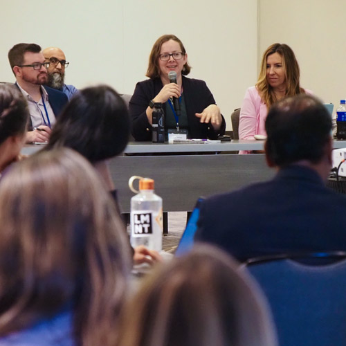 A person sitting at a u-shaped table speaking into a microphone with audience members watching