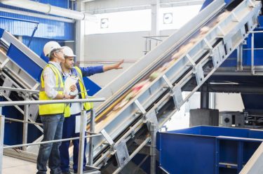 Two men in hard hats and safety vests stand talking and pointing inside recycling facility.