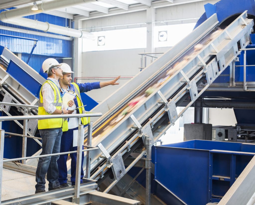 Two men in hard hats and safety vests stand talking and pointing inside recycling facility.