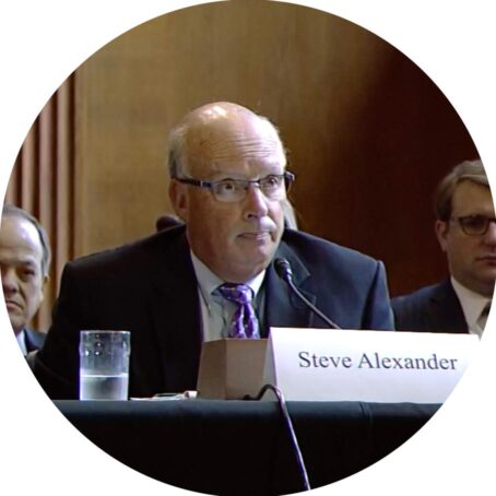 A man in a suit speaking into a microphone, with a plaque that reads 'Steve Alexander speaking on public policy