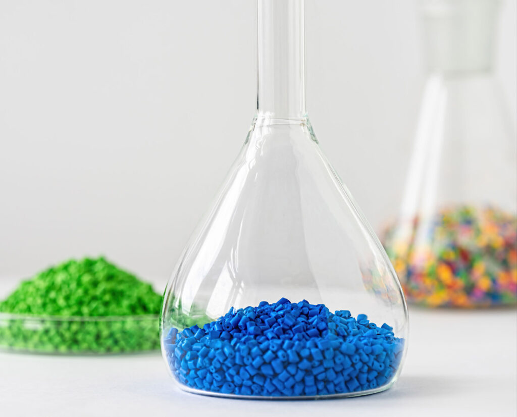 three lab flasks: one with bright green pellets, one with blue pellets, and one with multicolored pellets, all on a white surface.
