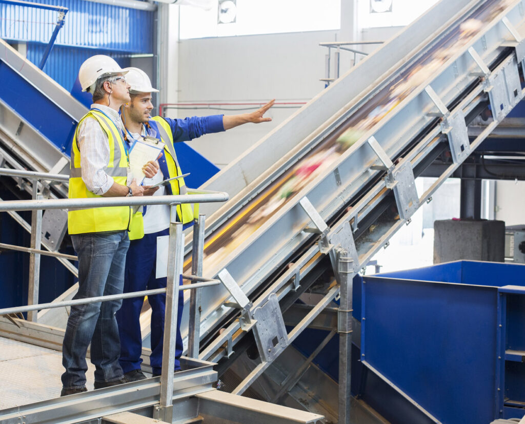 Two men in hard hats and safety vests stand talking and pointing inside recycling facility.